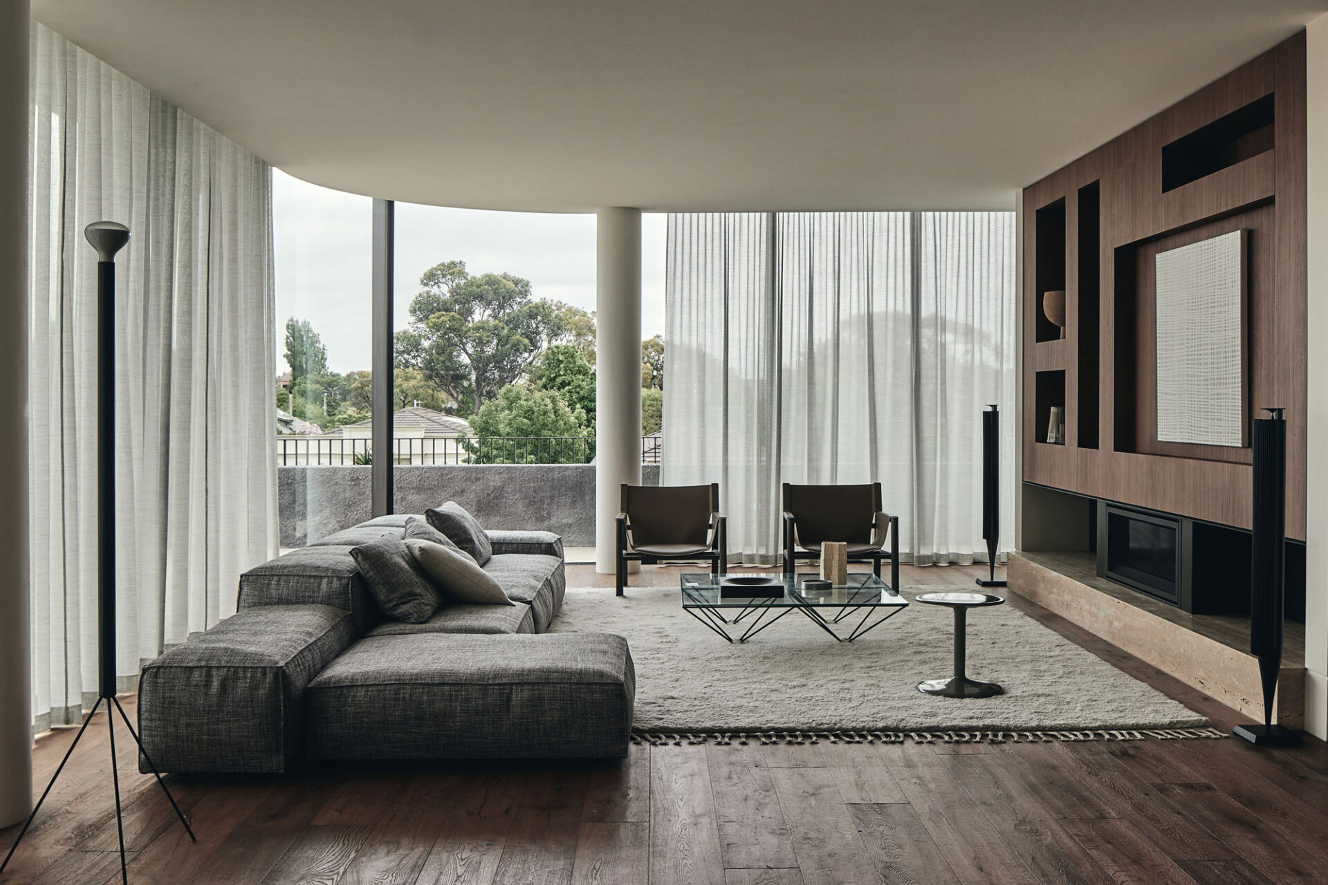 A spacious modern lounge room featuring curved glass window with sheer curtains framing the room. To the left is a large fabric sofa and to the right is wooden cabinetry with niches housing a collection of ceramics and artwork. In the centre of the room is a large light coloured rug with a glass coffee table and 2 occasional chairs.