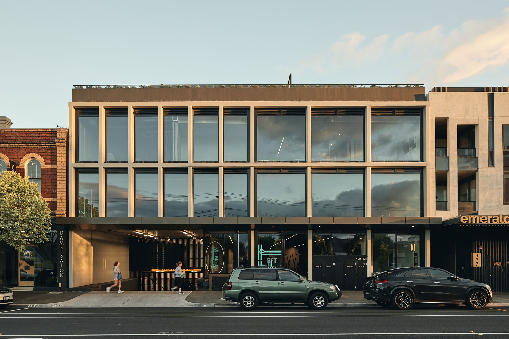 This building has a modern, boxy design with large glass windows framed by thin, light-coloured vertical and horizontal beams, creating a grid-like pattern. The ground floor has an open, recessed area with illuminated signage and entrances, while the upper floors feature reflective glass that mirrors the sky and surroundings. The exterior combines smooth surfaces with sharp lines, giving it a sleek and minimalist look.