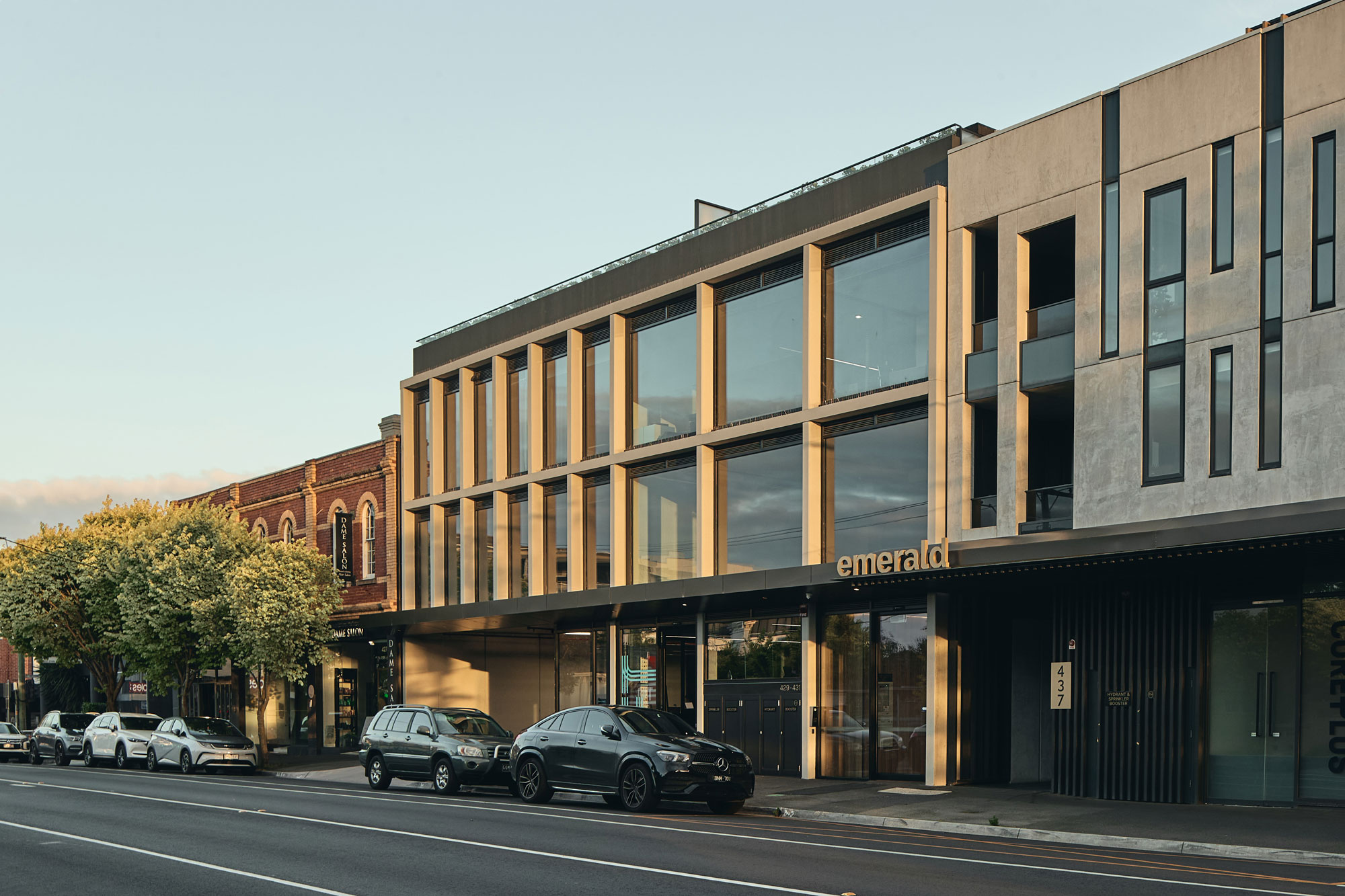 This building has a modern, boxy design with large glass windows framed by thin, light-coloured vertical and horizontal beams, creating a grid-like pattern. The ground floor has an open, recessed area with illuminated signage and entrances, while the upper floors feature reflective glass that mirrors the sky and surroundings. The exterior combines smooth surfaces with sharp lines, giving it a sleek and minimalist look.