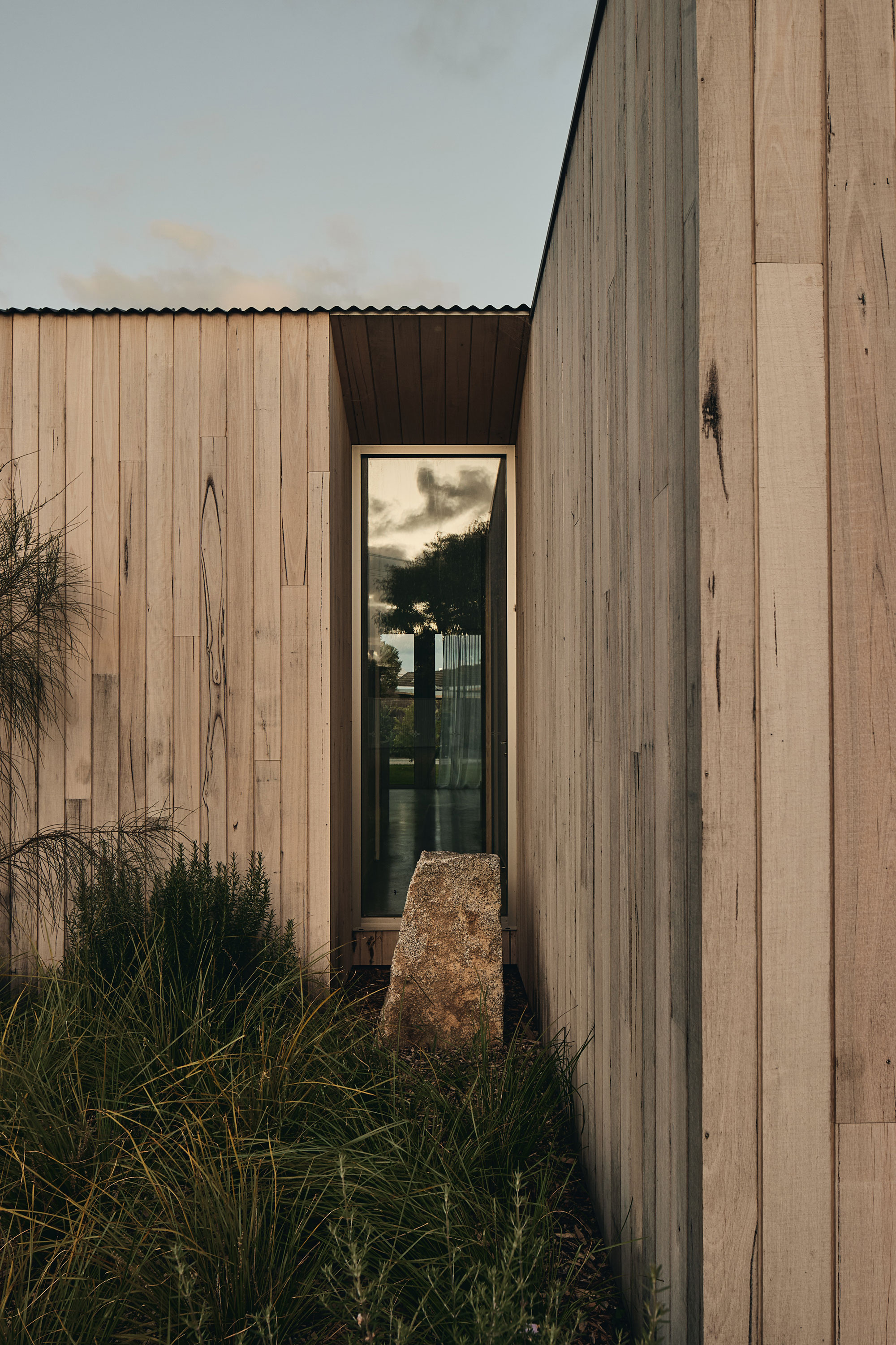 Soft morning light hits timber clad architectural house with flat roof and large boulder in front of a long feature window surrounded by lush native greenery.