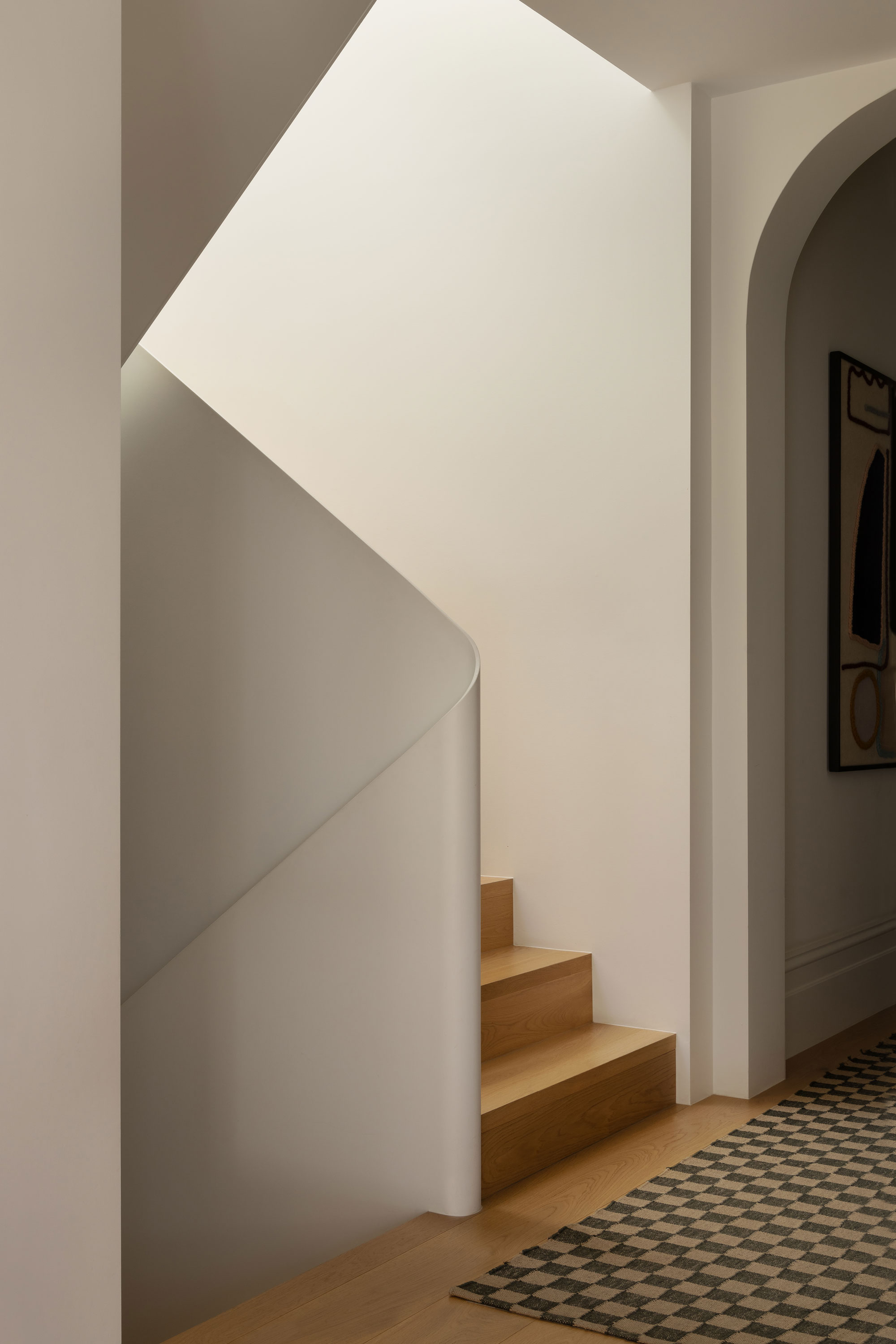 A sculptural staircase with soft light filtering from above., warm toned wood floors and a decorative carpet.