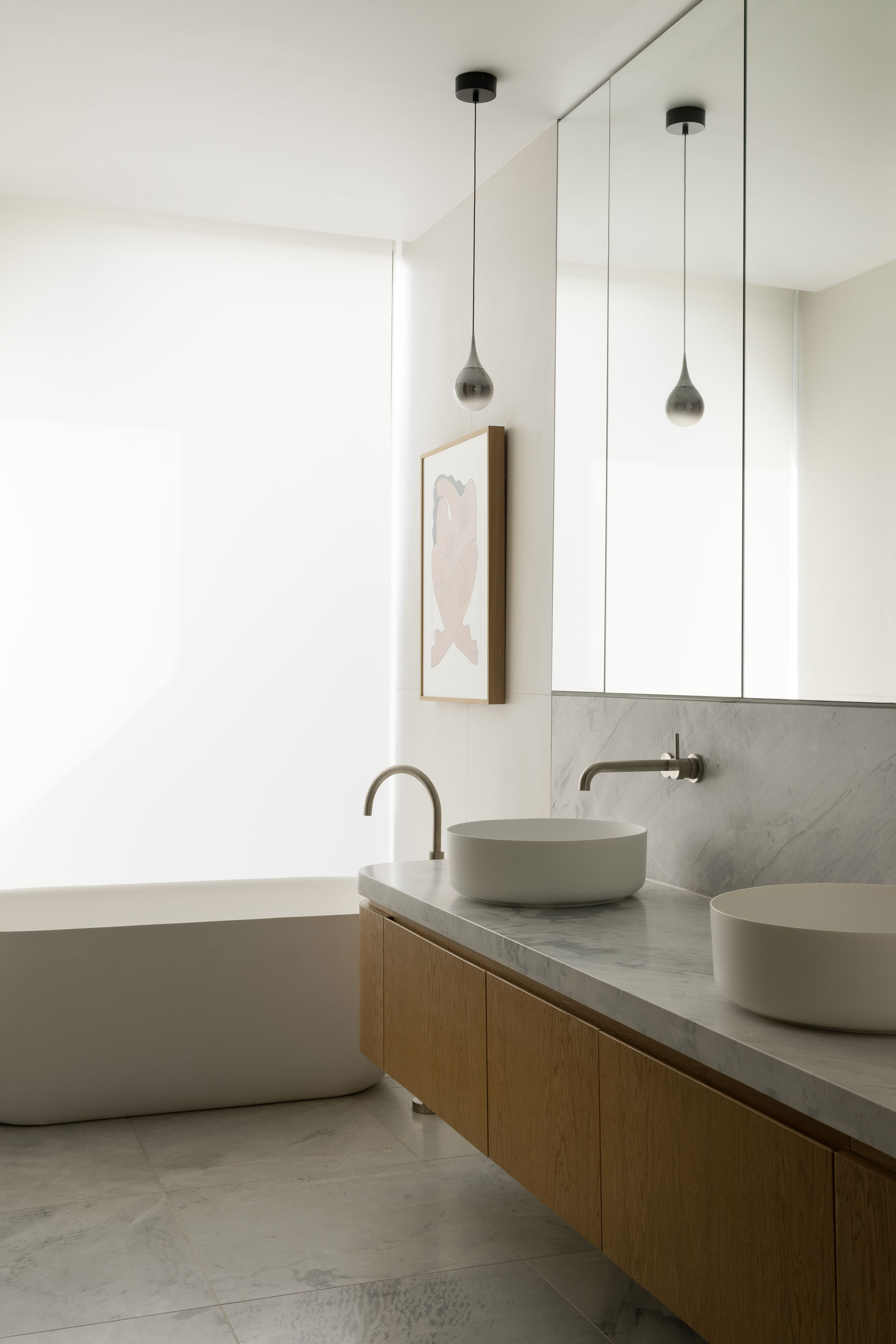 Soft light filters through a large window into a contemporary bathroom with stand alone bathtub, double vanity with wood cabinetry and a large mirror positioned above.