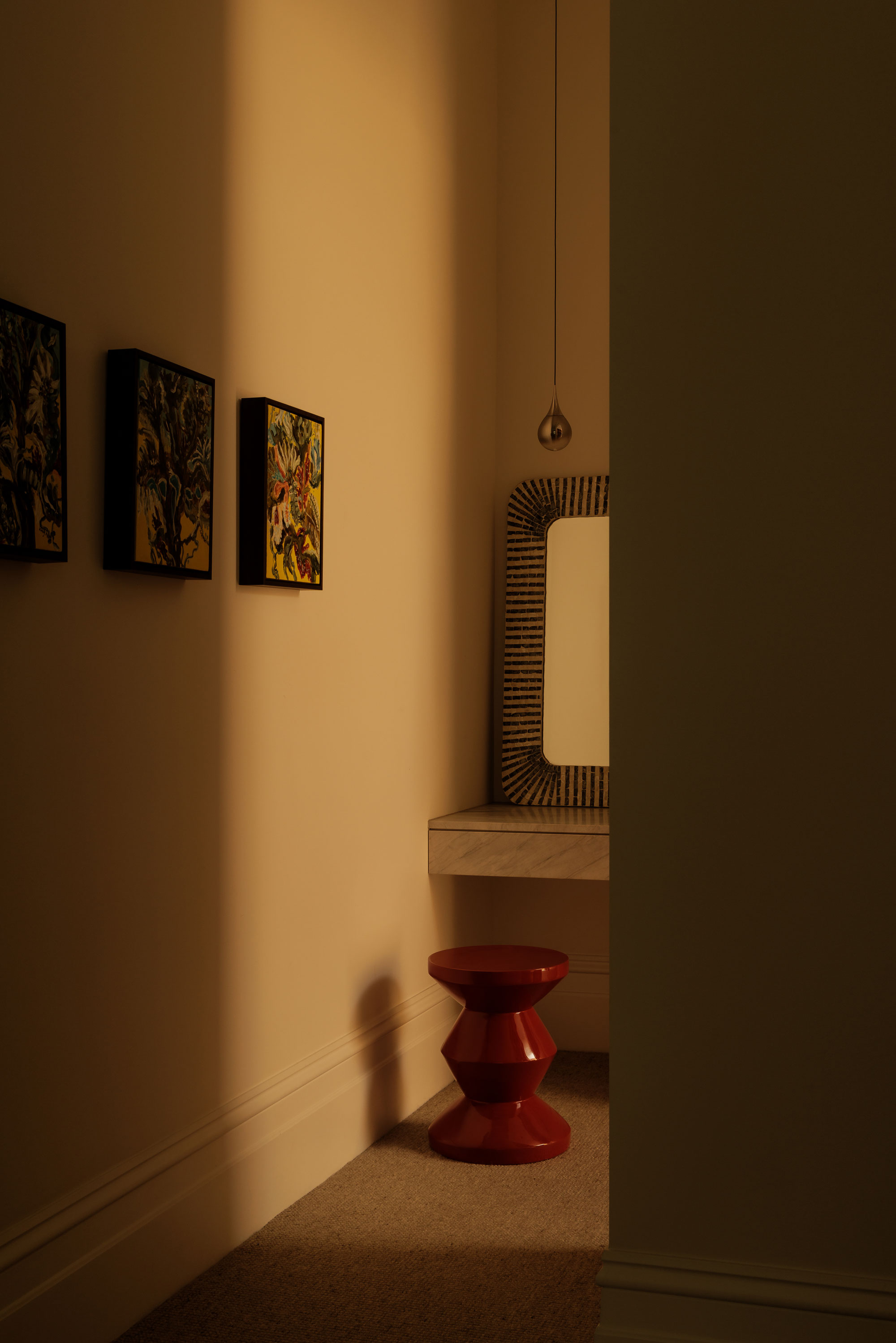 Warm light filters through dressing room, highlighting desk zone with red stool and mirror with pendant light hanging off centre and 3 small artworks.