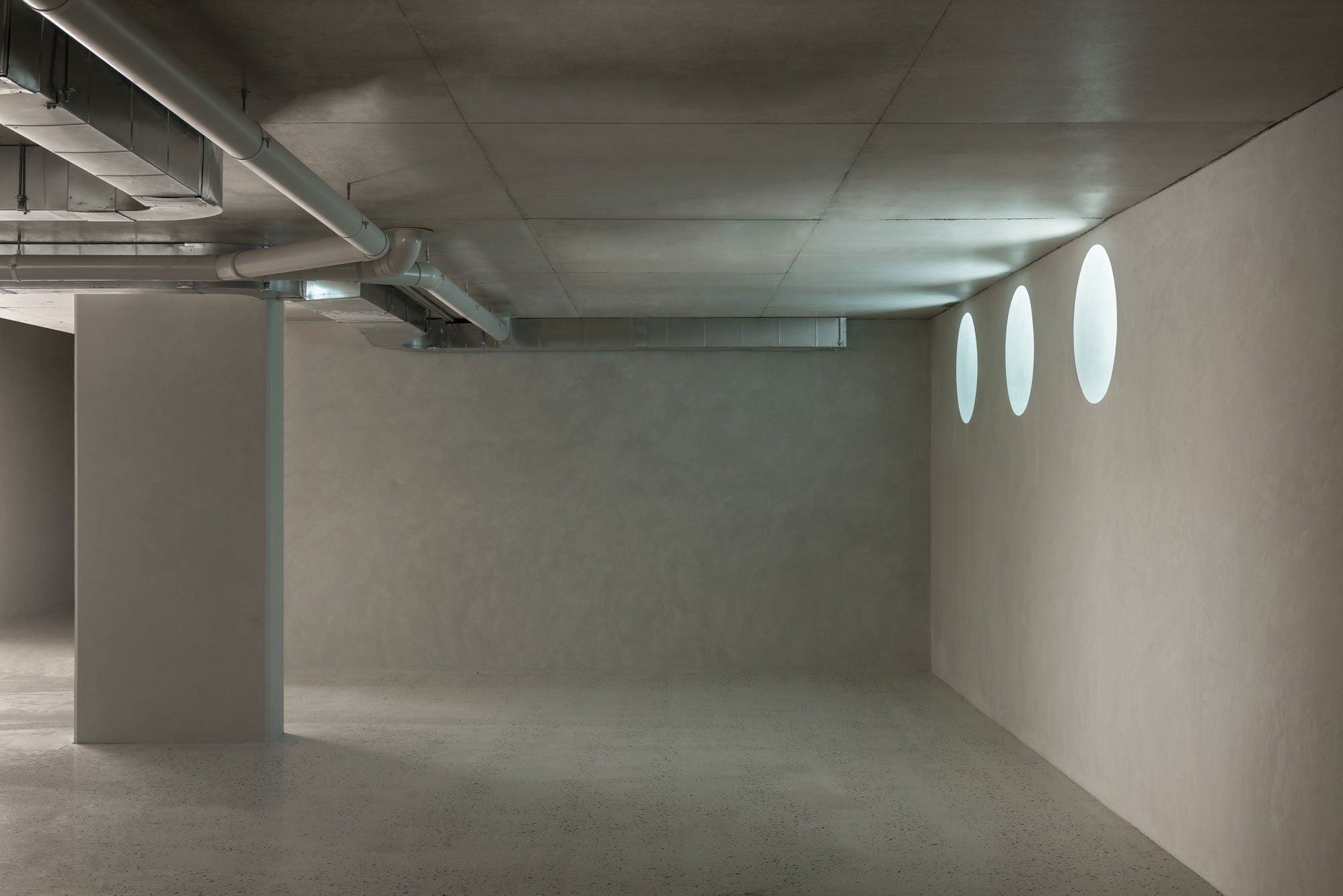 A spacious garage with concrete walls, polished concrete ceiling, exposed pipes and large column with three feature round windows.