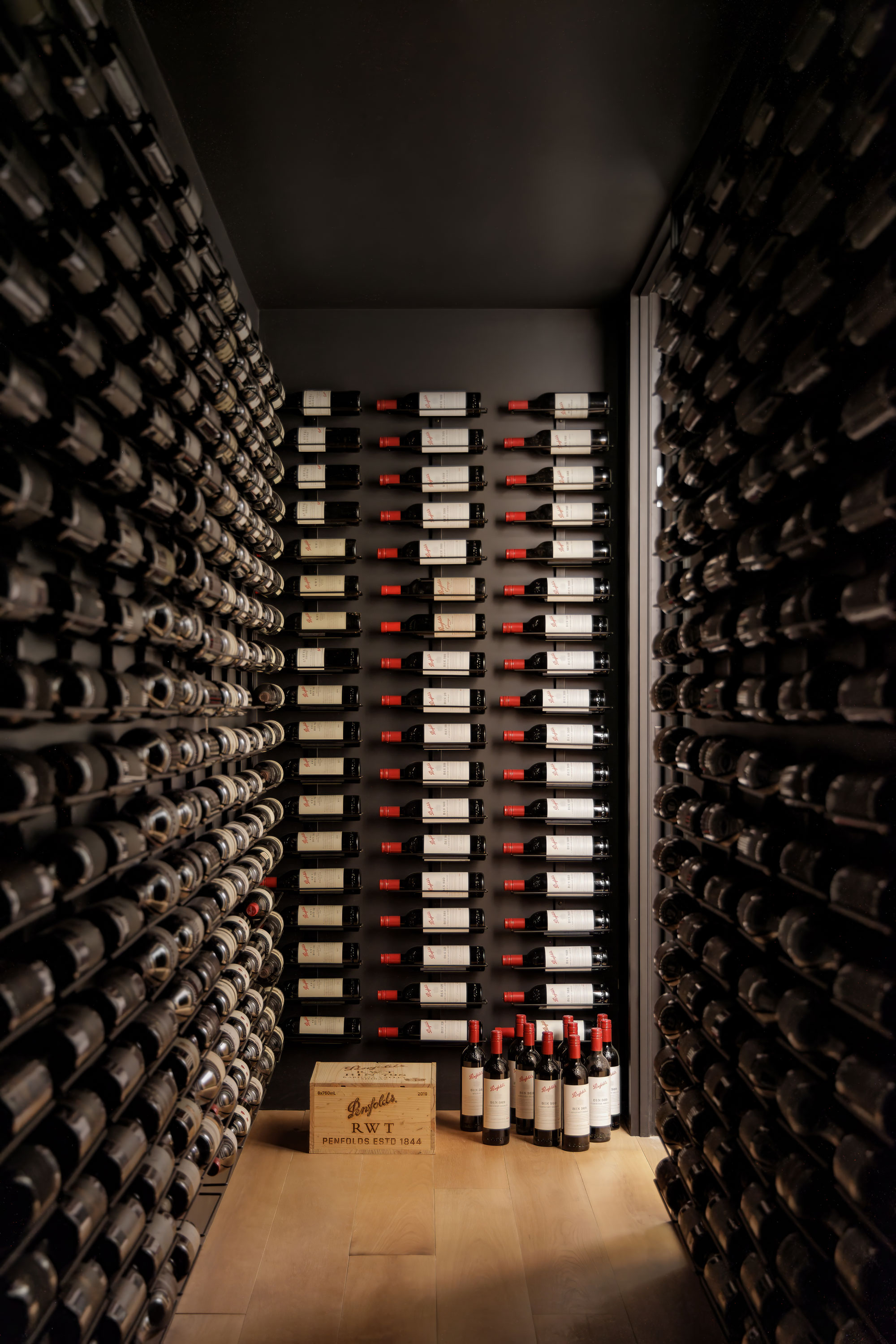 Long cellar with rows of wine complimented by dark walls and light coloured wood floors.