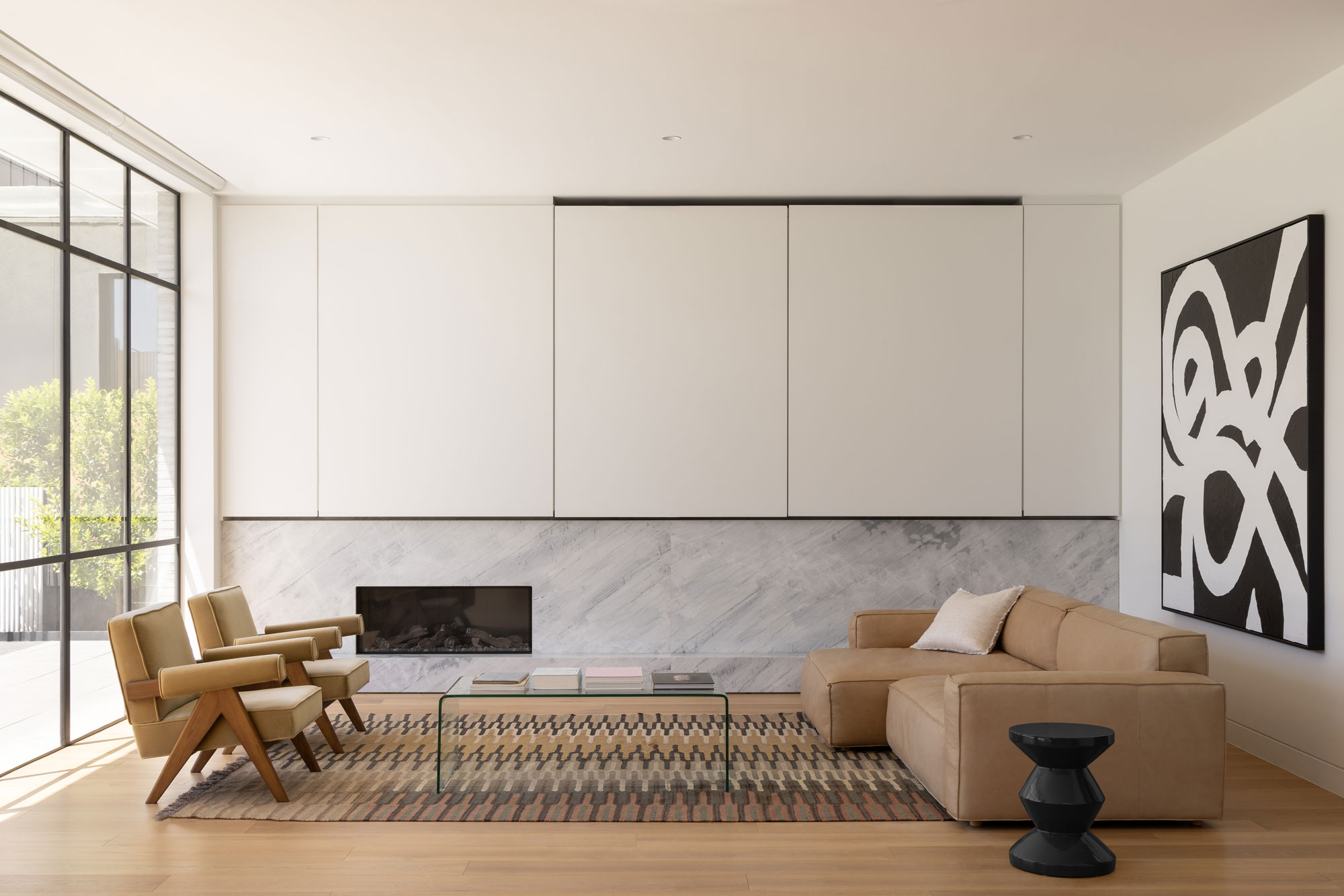 Large floor-to-ceiling windows with black frames open up into a modern lounge area with white walls, stone fireplace and two matching velvet chairs, stripe rug, large leather couch, glass coffee table and black & white abstract artwork.