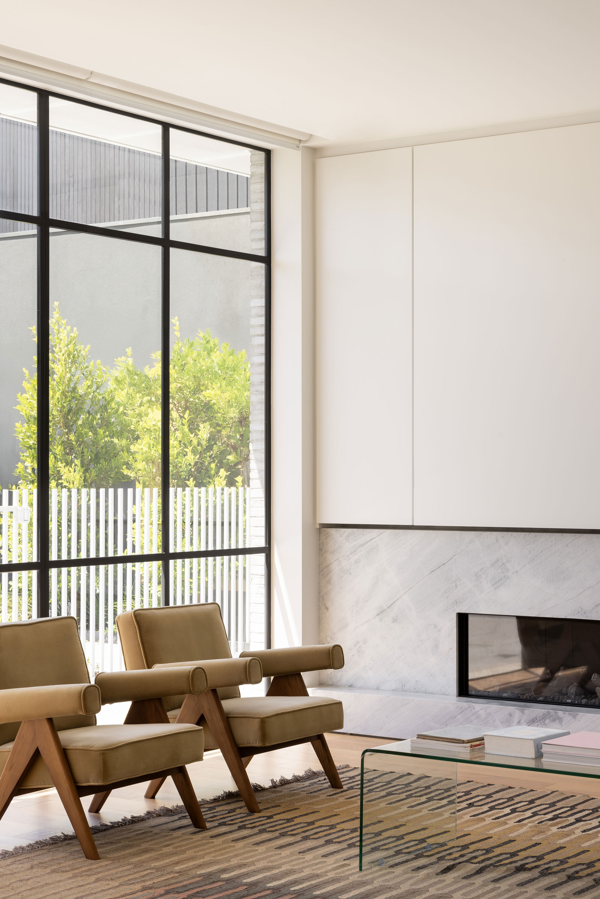 Large floor-to-ceiling windows with black frames open up into a modern lounge area with white walls, stone fireplace and two matching velvet chairs, stripe rug and glass coffee table.