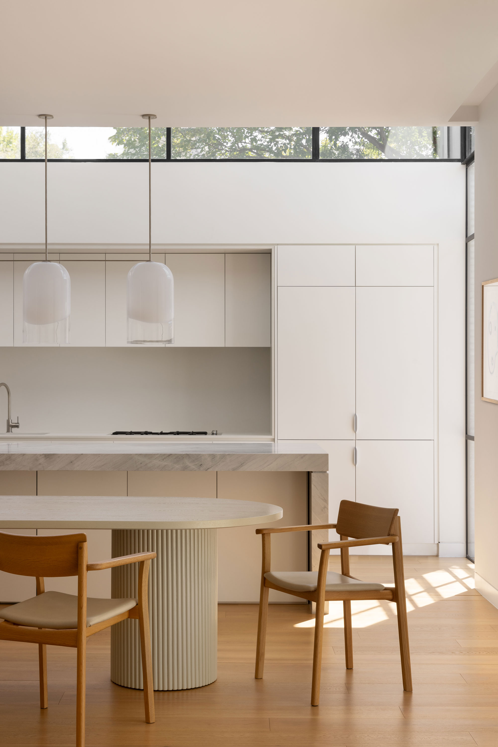 The space has light wooden flooring and white cabinetry that blends seamlessly into the walls. A marble countertop extends over the kitchen island, adding a touch of elegance. Two large, delicate glass pendant lights hang from the ceiling, providing soft illumination. In the foreground, there is a round dining table with a fluted, textured base, accompanied by two wooden chairs with a warm, natural finish. Sunlight streams in from high windows, casting soft shadows and highlighting the airy and peaceful atmosphere of the room.