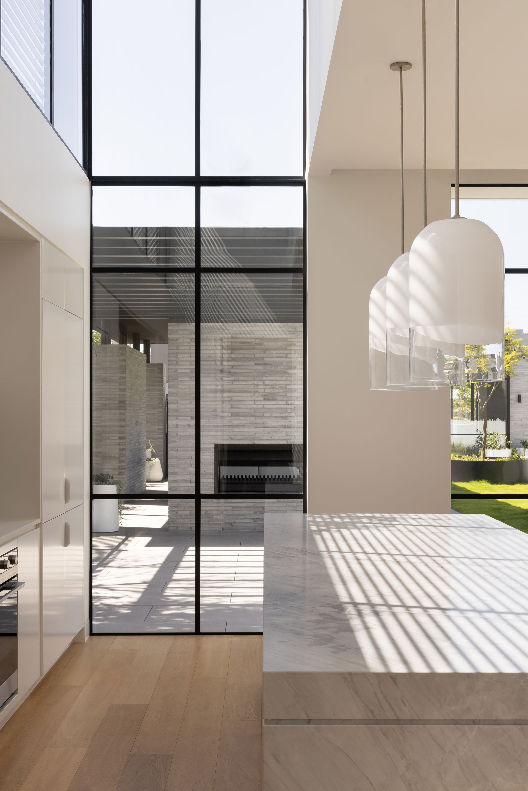 The space has light wooden flooring and white cabinetry, large floor-to-ceiling windows with black frames and a marble countertop that extends over the kitchen island. Two large, delicate glass pendant lights hang from the ceiling, providing soft illumination. Sunlight streams in from high windows, casting soft shadows and highlighting the airy and peaceful atmosphere of the room.