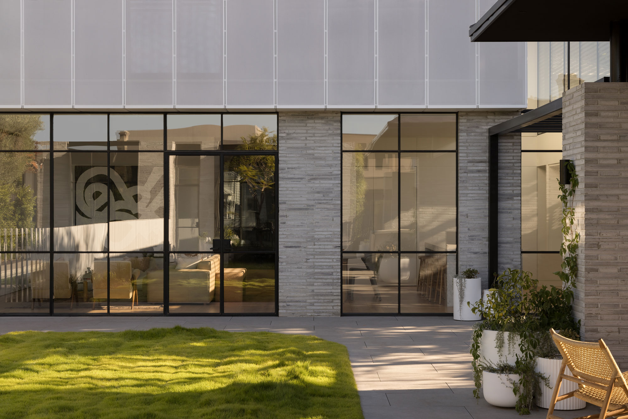 Soft light filters through garden casting shadows on lush lawn in front of a modern double-storey home with large floor to ceiling windows, brick work and metal façade detailing.