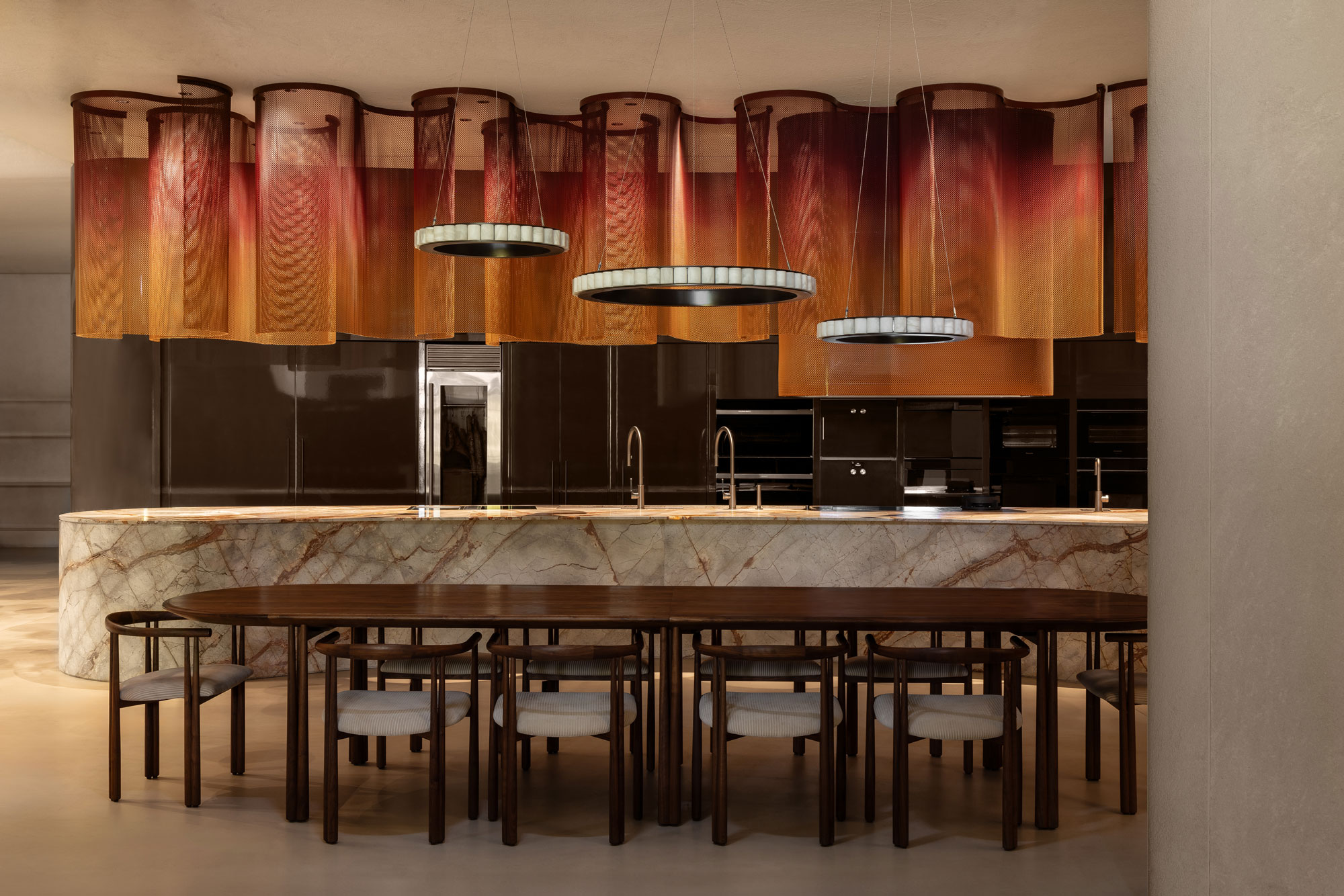 Landscape image of luxurious modern kitchen with sculptural lighting fixtures, large marble island bench set behind a long line dining table with matching seating.