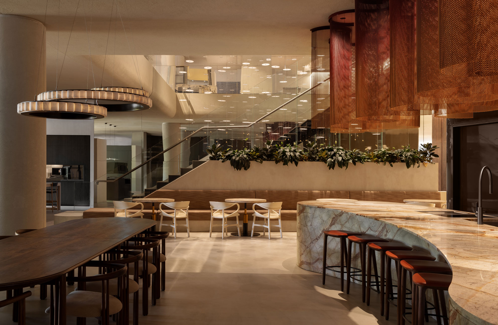 Landscape image of luxurious modern kitchen with sculptural lighting fixtures, large marble island bench set behind a long line dining table with matching seating.