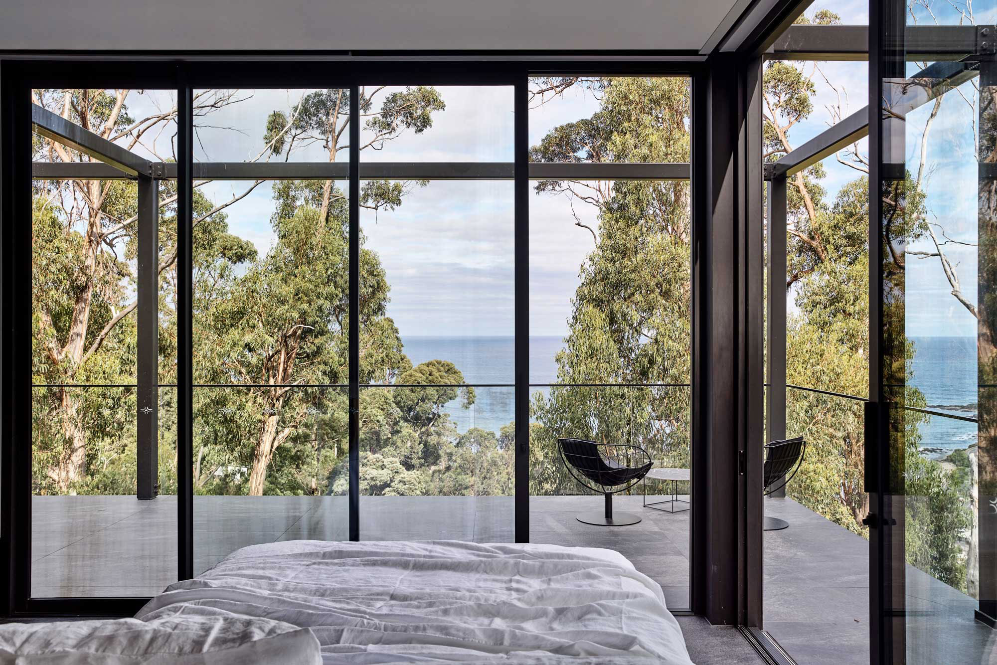 A bed looking out to panoramic views of the ocean and lush greenery, framed by floor to ceiling black metal doors that open onto a large balcony area.