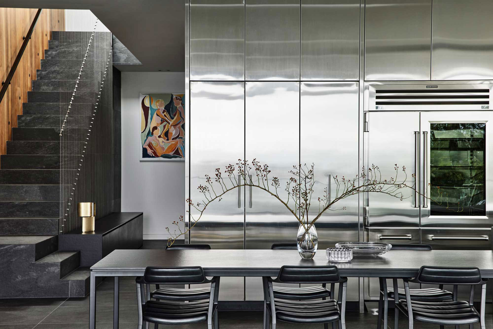 Modern stainless steel kitchen with sleek black furnishings, dark concrete flooring, warm timber panelled walls and staircase to the left.