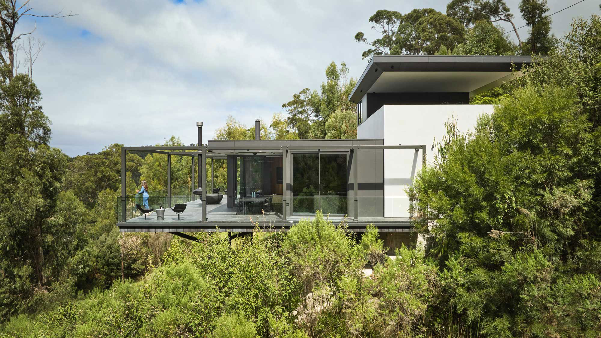 House perched in the tree tops with large balcony with woman looking out to the view.