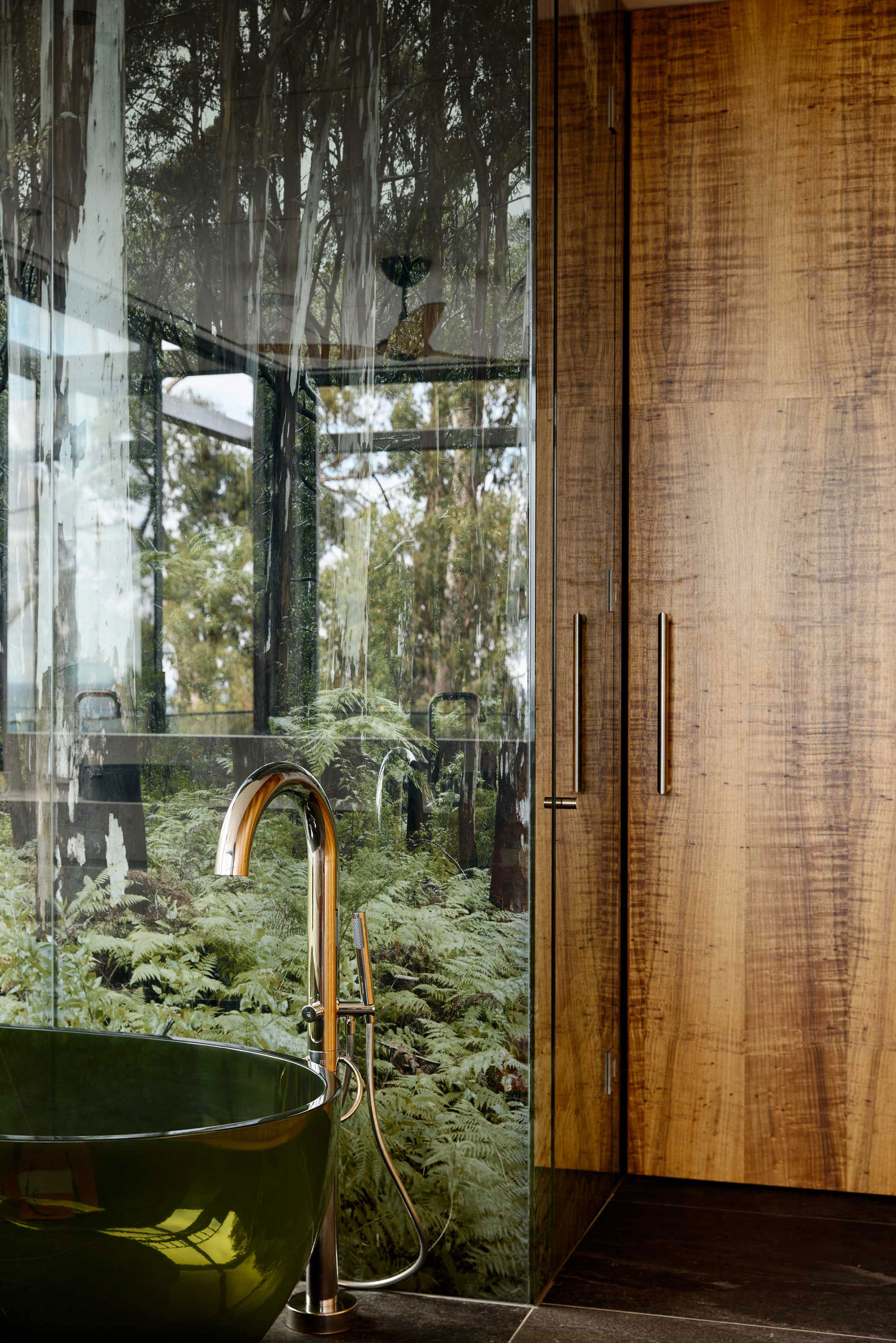 Moody bathroom with large timber door, green bathtub and chrome faucet and custom wallpaper.