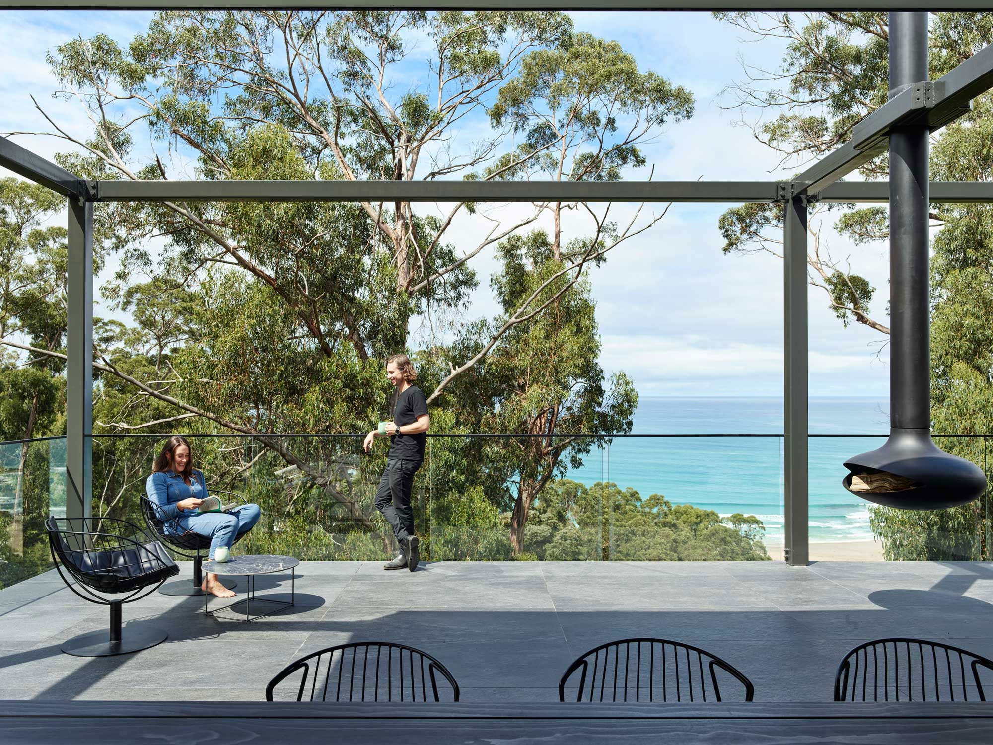 Two people are on a patio with glass railings, overlooking trees and the ocean. One person is seated, the other is standing. A hanging fireplace is in the foreground.