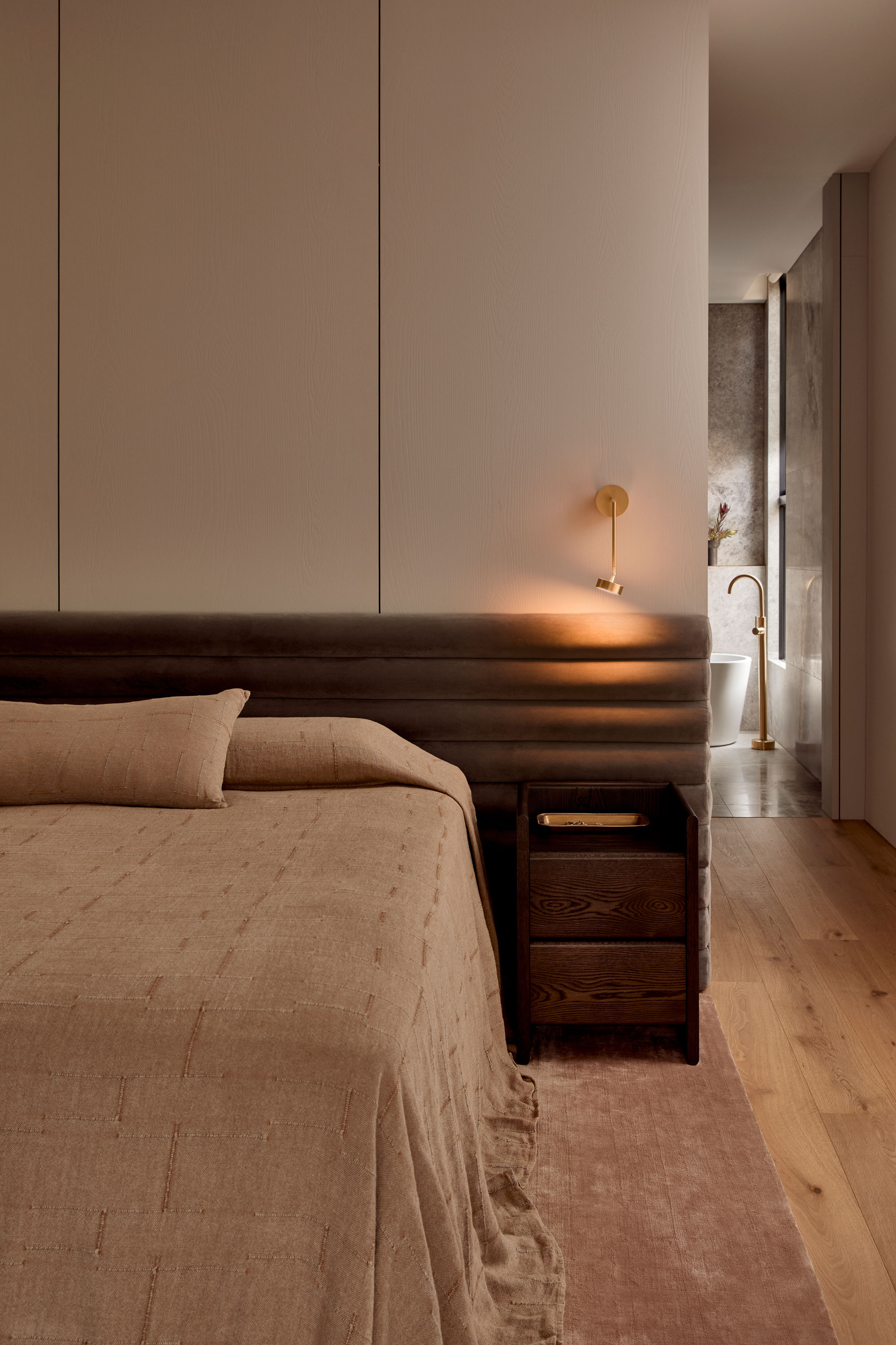 Minimalist bedroom with taupe bedding, velvet headboard, oak bedside table, and brass wall light, leading to a stone-clad bathroom with a freestanding tub and brass fixtures.