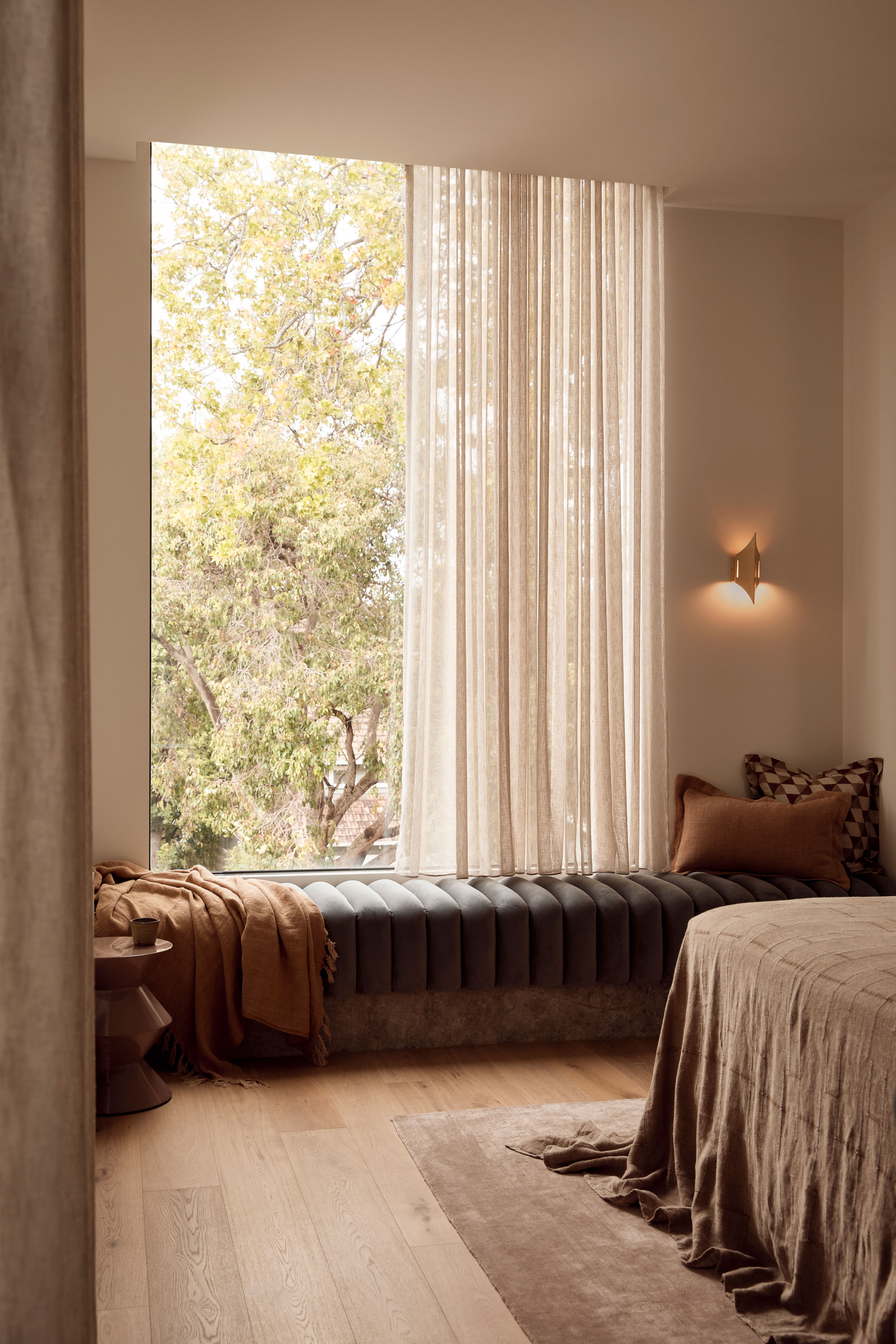 Bedroom corner with a ribbed velvet window seat, sheer curtains, textured bedding, and soft timber flooring, framed by a large window with views of leafy trees outside.