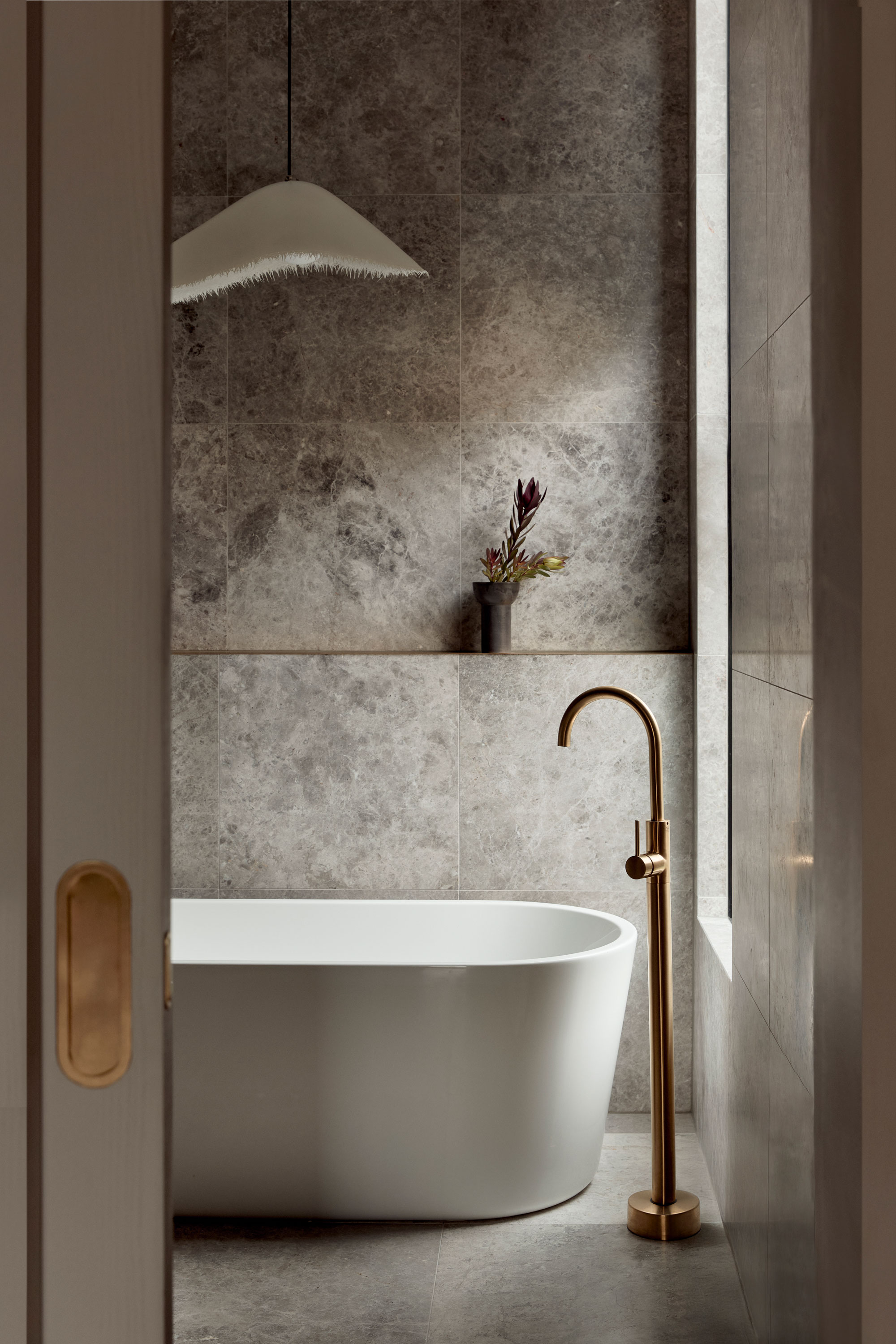 Modern bathroom with a white freestanding tub, brushed brass floor-mounted tap, grey stone tile walls, and a sculptural pendant light above, with a single vase on a recessed shelf.