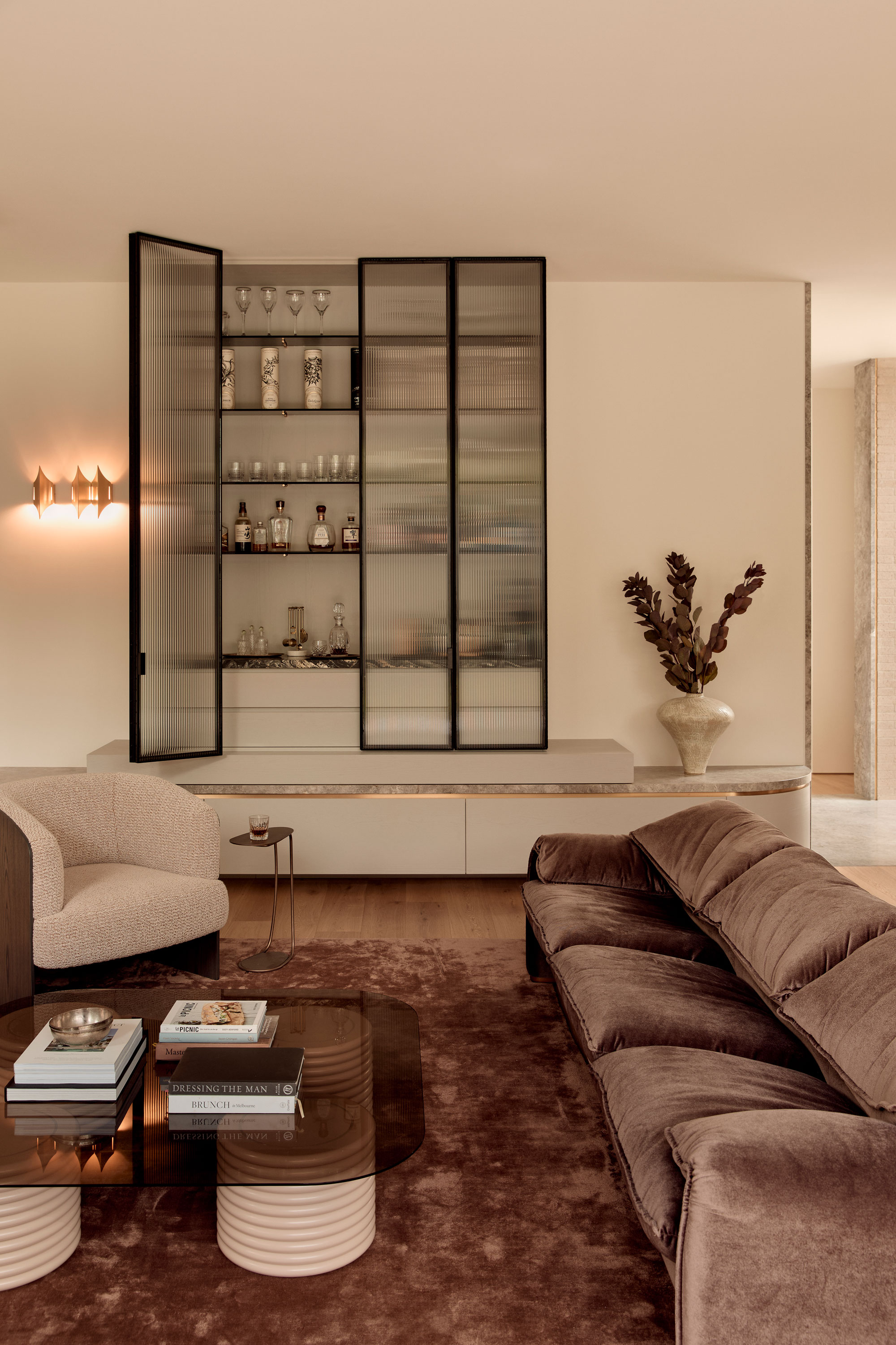 Contemporary open-plan living and dining room with velvet sofa, built-in bar with fluted glass and black metal framing, marble accents, timber floors, minimalist decor, modern sculptural furniture, and soft ambient lighting in neutral tones.