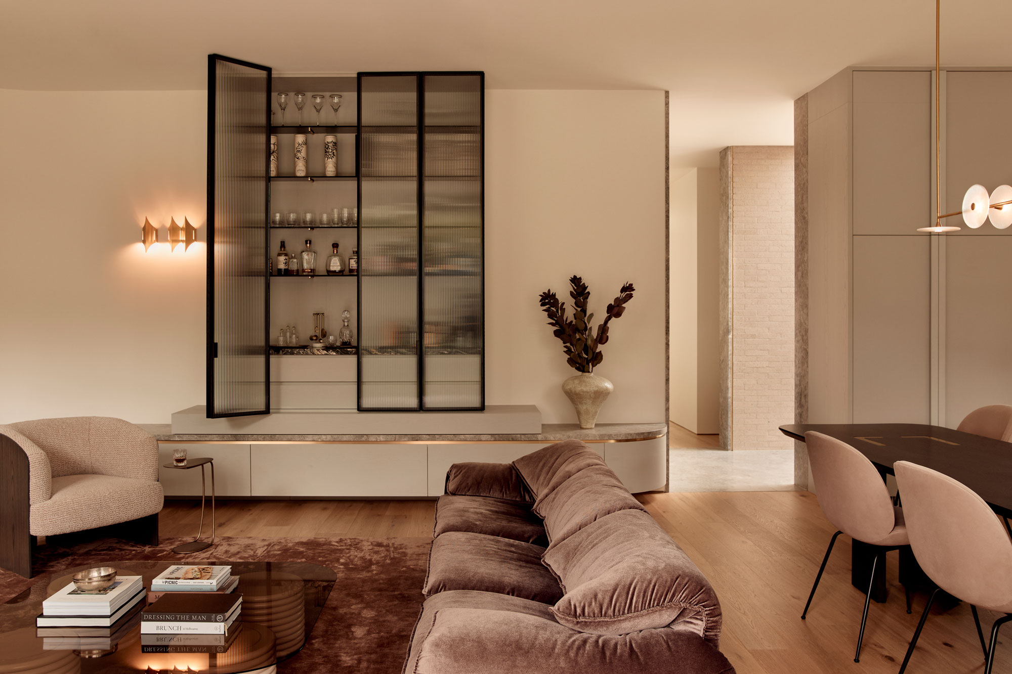 Contemporary open-plan living and dining room with velvet sofa, built-in bar with fluted glass and black metal framing, marble accents, timber floors, minimalist decor, modern sculptural furniture, and soft ambient lighting in neutral tones.