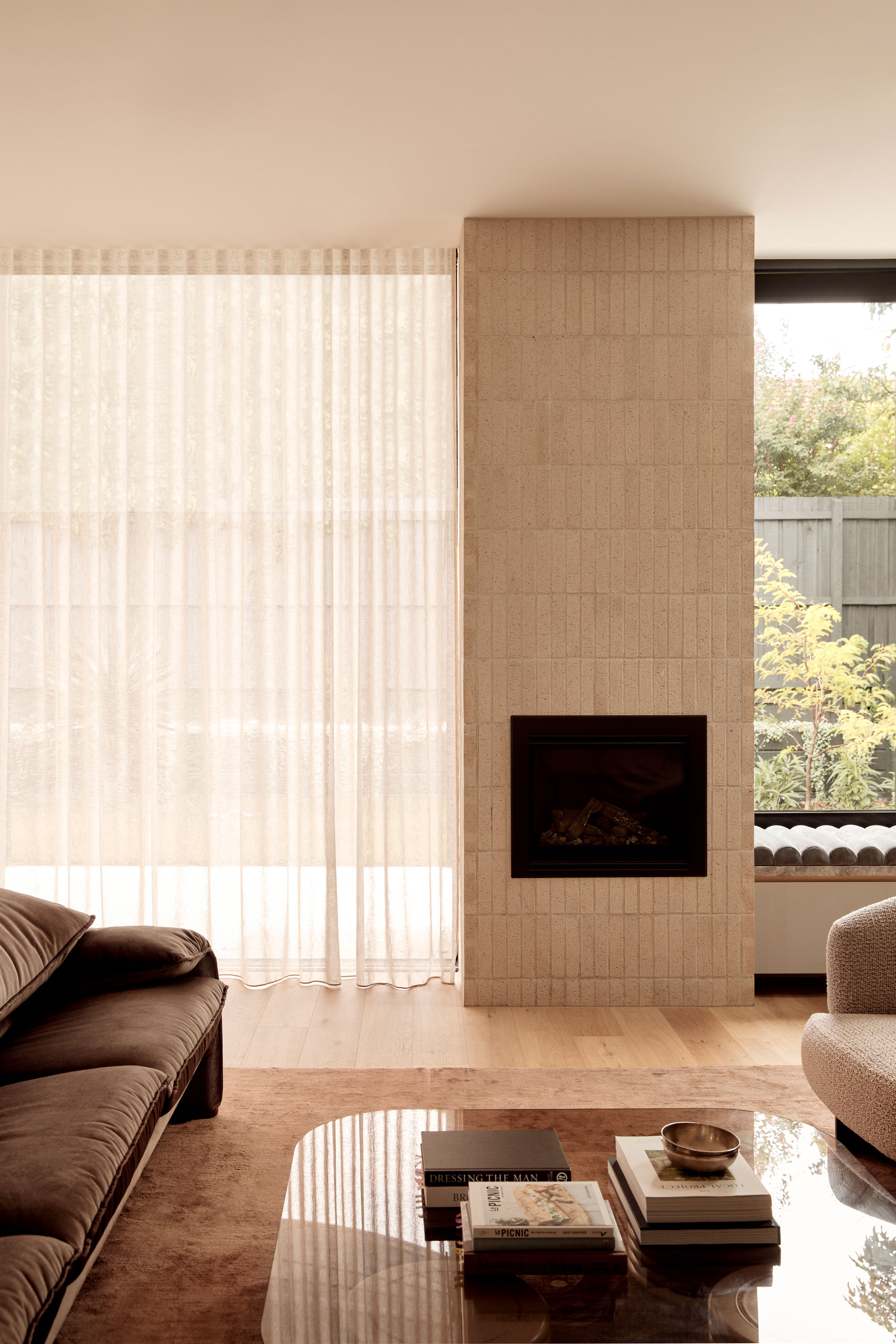 Modern light-toned brick fireplace centred around a lounge room with lush velvet furnishings, sheer curtains and glass coffee table. A large window off centre looks out onto green landscaping.
