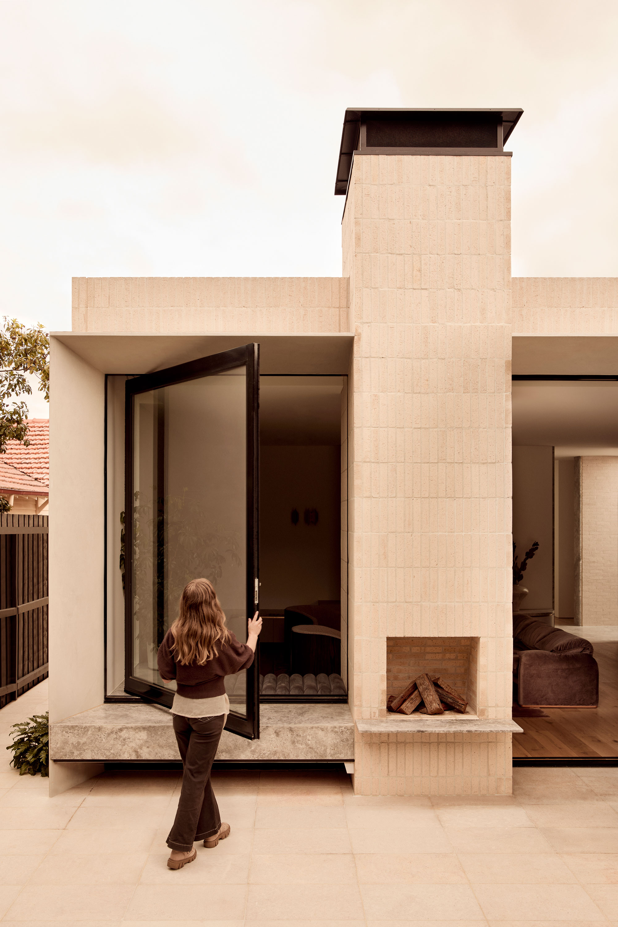 Modern light-toned brick outdoor fireplace with large pivot window opening out to a stone bench seat.