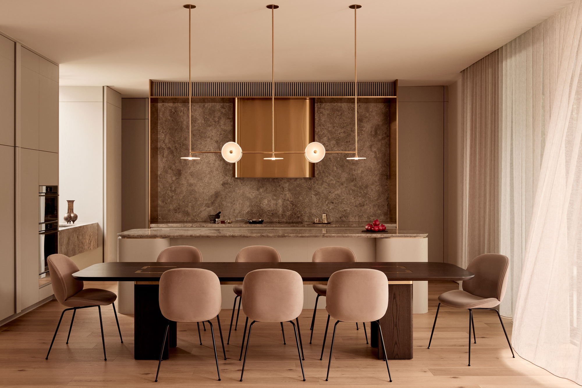 Minimalist open-plan kitchen and dining area featuring a dark wood table with eight blush velvet chairs, a stone marble island and splashback, brass pendant lights, and soft natural light filtering through sheer curtains.