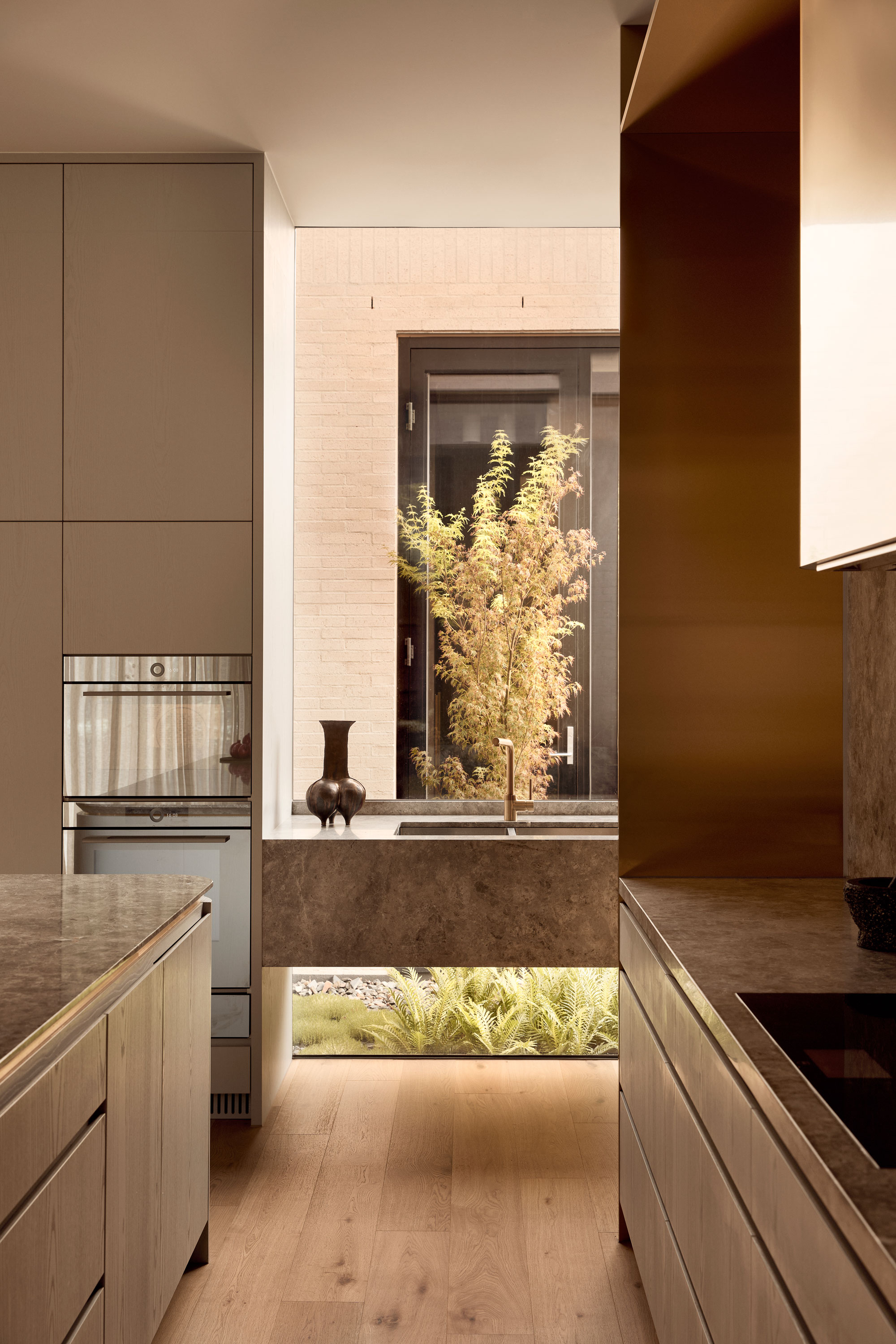 Minimalist open-plan kitchen featuring a large floor to ceiling window, looking out to landscaping, a stone marble island and splashback, brass range hood, and soft natural light filtering through.