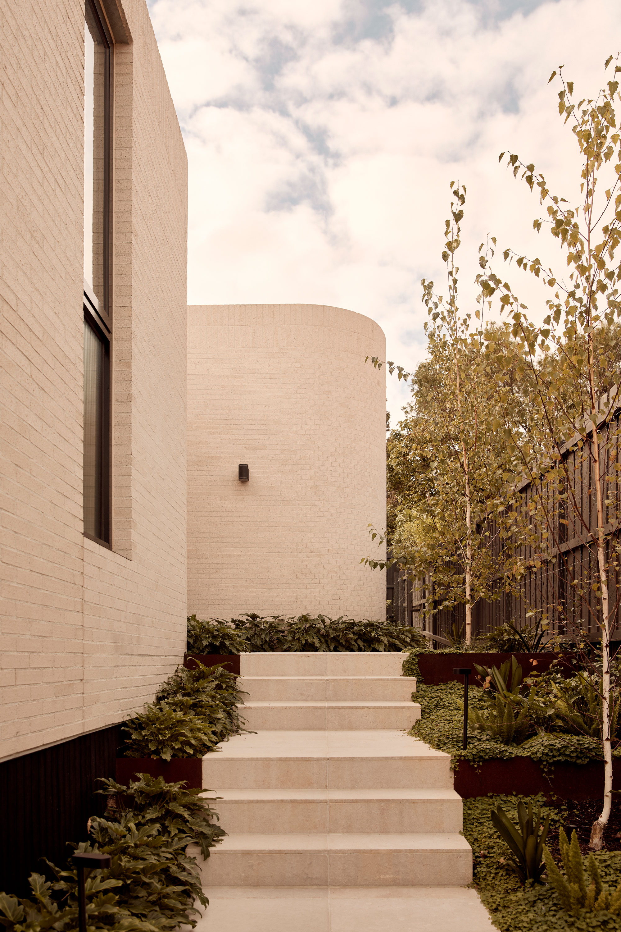 Modern two-storey house with a minimalist design, featuring a light-toned brick staircase that leads to a curved brick wall. Lush landscaping frames the walkway.