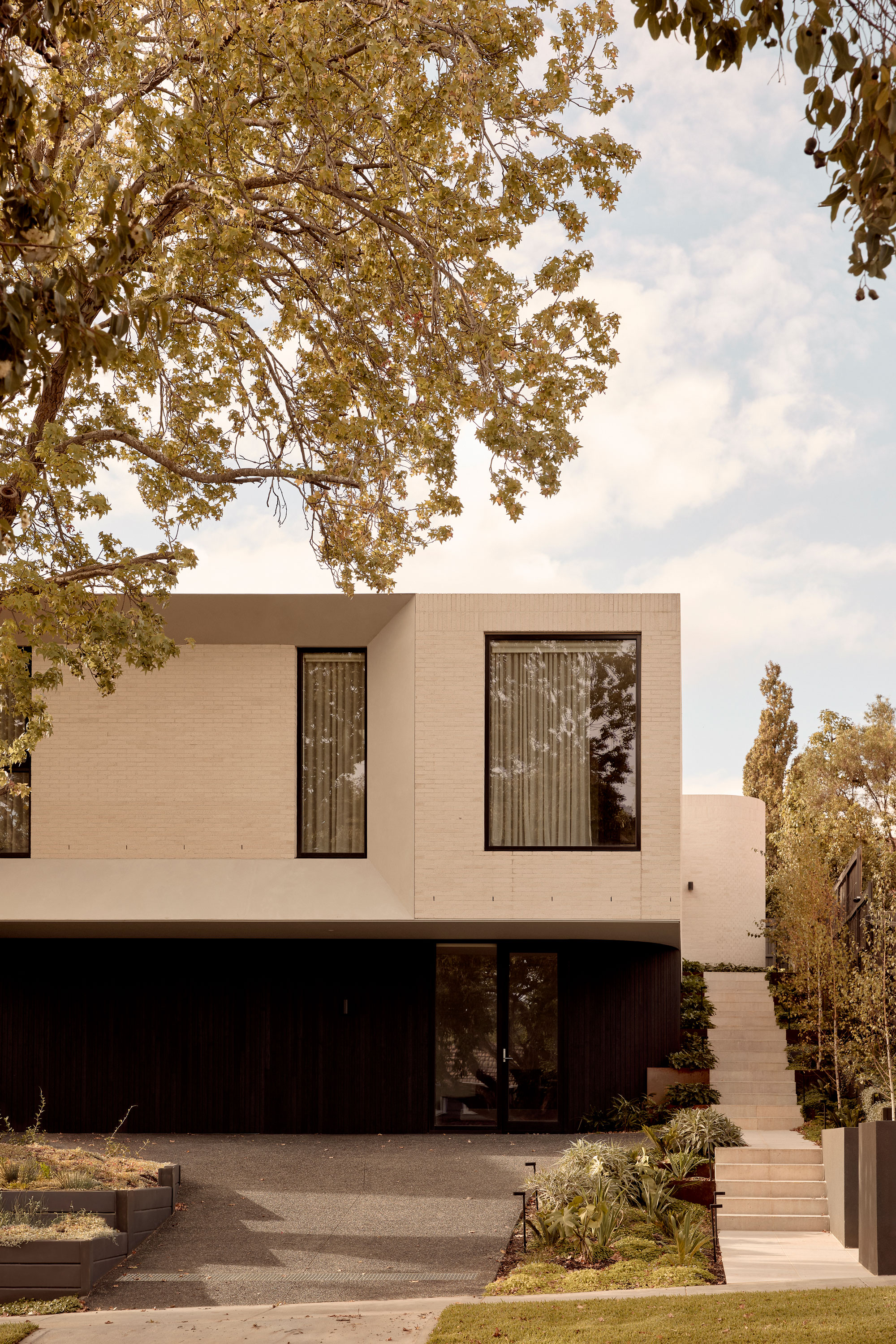 Modern two-storey house with a minimalist design, featuring a light-toned brick upper level that cantilevers over a dark timber-clad ground floor. The façade includes large vertical windows with sheer curtains, a shadowed driveway entry, and a landscaped side stair framed by soft greenery.
