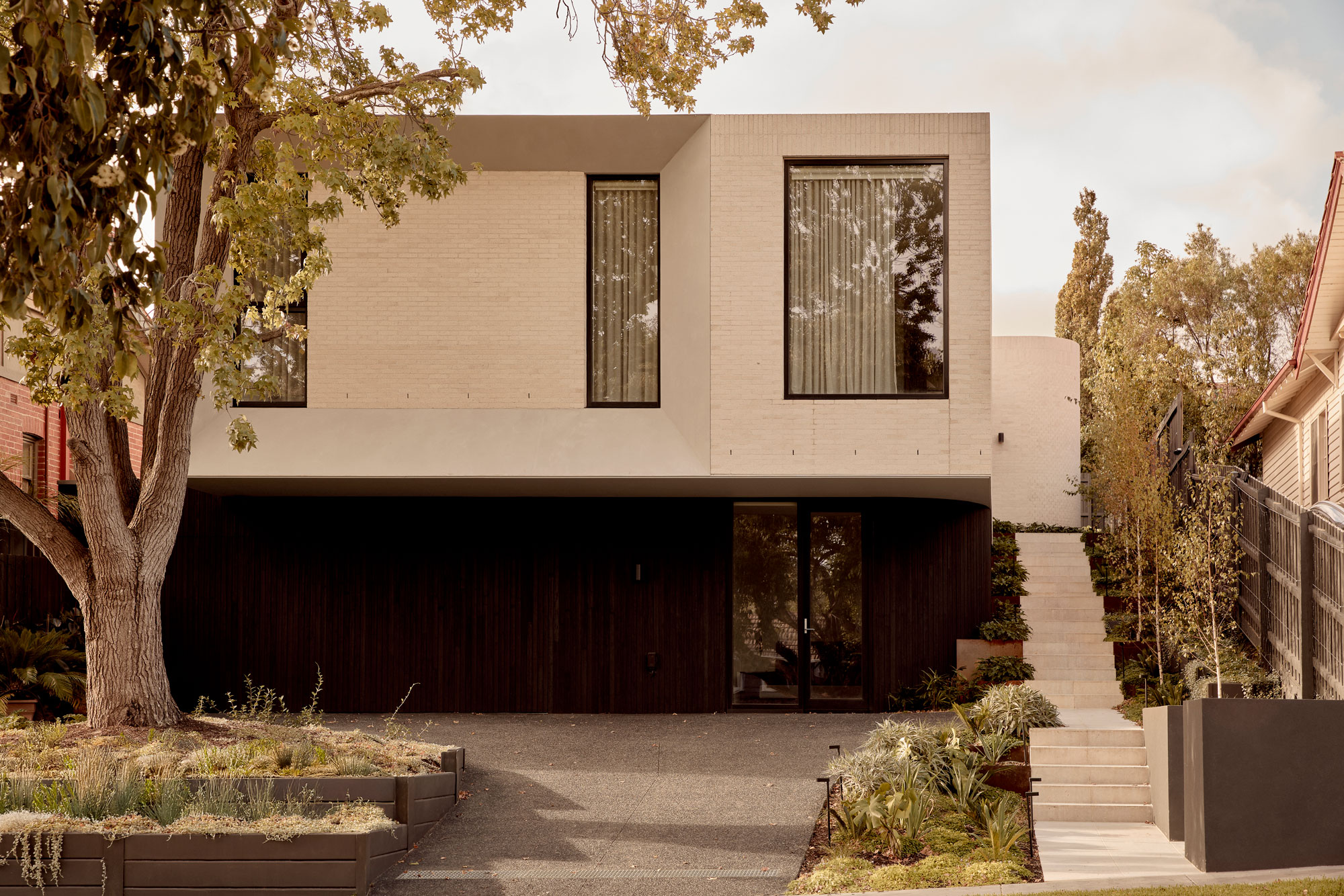 Modern two-storey house with a minimalist design, featuring a light-toned brick upper level that cantilevers over a dark timber-clad ground floor. The façade includes large vertical windows with sheer curtains, a shadowed driveway entry, and a landscaped side stair framed by soft greenery.